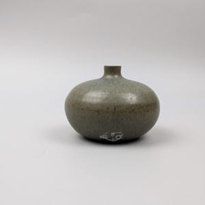 May include: A small, round, ceramic vase with a short neck. The vase has a speckled, muted green and brown glaze. The base of the vase has a small, decorative design. The vase is a decorative home decor item.