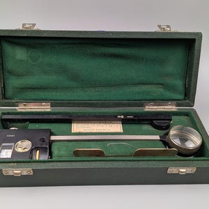 May include: A dark green case with a green velvet lining contains a black and silver precision instrument. The instrument features a magnifying glass, a ruler, and a small level. The case is secured with metal clasps.