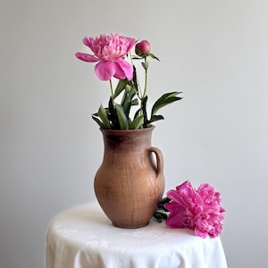 May include: A brown ceramic pitcher with a single handle containing a bouquet of vibrant pink peony flowers and a closed bud, with an additional fully bloomed pink peony resting beside the base of the pitcher on a white surface.