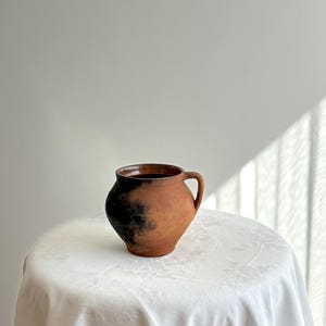 Puede incluir: Una jarra de cerámica de color terracota con un asa. La jarra tiene un cuerpo redondeado y un patrón oscuro y moteado cerca de la base. Se encuentra sobre un mantel blanco texturizado, lo que sugiere una estética vintage o artesanal.