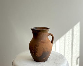 May include: A rustic, brown earthenware jug with a handle, set on a white tablecloth. The jug has a wide base, a rounded body, and a dark rim. The surface shows variations in color, with darker patches. The jug is likely vintage or antique.