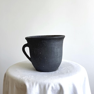 Puede incluir: Una taza de cerámica negra mate con una superficie texturizada que presenta líneas verticales y una banda decorativa de patrones geométricos cerca del borde. La taza tiene un asa curva en el lado izquierdo y una abertura ancha y acampanada.