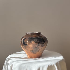 Puede incluir: Un jarrón de cerámica de color terracota con un cuerpo ancho y un cuello estrecho. El jarrón tiene marcas oscuras y moteadas cerca de la parte superior y se exhibe sobre una tela blanca. El jarrón es probablemente hecho a mano y adecuado para la decoración del hogar.