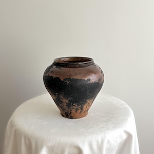 May include: A vintage pottery vase with a rounded body and a narrow base. The vase has a dark brown rim and a black and brown exterior, giving it a weathered look. It is displayed on a white tablecloth.