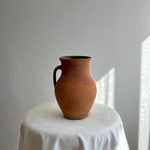 Könnte beinhalten: Ein rustikaler Krug aus Terrakotta mit einem Griff. Das irdene Gefäß hat einen abgerundeten Körper und einen leicht ausgestellten Rand. Er steht auf einer weißen, drapierten Oberfläche.