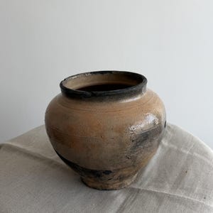 May include: A rustic pottery vase with a rounded body and a dark, slightly flared rim. The vase has a mottled, earthy colour palette with shades of tan, brown, and black, suggesting a natural, aged aesthetic. The vase is displayed on a neutral-toned linen cloth.