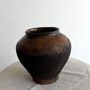 May include: A rustic, handmade ceramic vase with a rounded body and a dark brown rim. The vase features a gradient of colors, transitioning from a dark, almost black base to a lighter brown and orange near the top. The vase sits on a neutral-colored cloth.