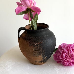 Peut inclure: Une cruche en céramique ronde et rustique aux tons sombres avec une anse unique, présentant une surface texturée et patinée passant du noir au brun terreux. Une pivoine rose vif avec des feuilles vertes est placée dans la cruche.