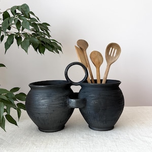May include: A double-chambered black ceramic utensil holder featuring a central circular handle connecting two rounded pots with textured, etched surfaces. Several light-colored wooden kitchen spoons and a spatula are placed inside.
