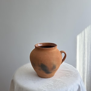 May include: A terracotta-coloured, handmade ceramic jug with a single handle. The jug has a wide, rounded body and a slightly flared rim. Darker markings are visible on the jug's surface. The jug is displayed on a white, patterned tablecloth.