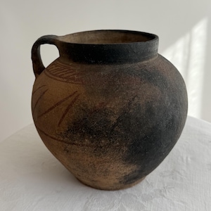Op de afbeelding: Een rustieke, handgemaakte keramische kruik met een afgeronde body en een enkele handgreep. De kruik heeft een donkerbruin en zwart kleurenschema met geëtste patronen in de buurt van de bovenkant. De kruik wordt waarschijnlijk gebruikt voor decoratieve doeleinden.