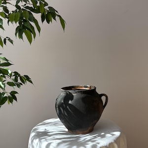 May include: A dark, rustic ceramic pot with a single handle. The pot is a deep black colour with hints of brown and sits on a white cloth. Green leaves are visible in the upper left corner, casting shadows on the pot.