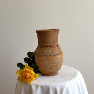 May include: A rustic, light brown terracotta pitcher with a textured, porous surface and a flared rim. The vessel features two horizontal dark brown bands encircling the midsection, with a single yellow rose placed at its base.