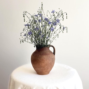 Puede incluir: Una jarra de arcilla marrón, redonda y rústica, con una sola asa, llena de un ramo de delicadas flores de lino azul de tallo fino. La jarra tiene una superficie texturizada y desgastada, con un cuerpo ancho y redondeado que se estrecha.