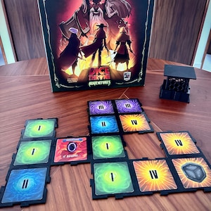以下が含まれることがあります： 角のある人物が描かれたDice Throne Adventuresボードゲームの箱、黒いプラスチック製のダイスタワー、テーブル上に配置された正方形のゲームタイル。タイルにはローマ数字のI、II、III、IVと、It Beginsと書かれたCrimson Sandsカードが表示されています。