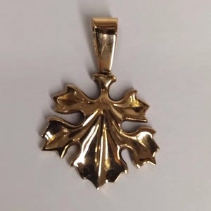 May include: A polished gold-tone pendant shaped like a stylized maple leaf with a smooth, reflective surface and a vertical bail at the top for attachment to a chain. The metal has a warm, metallic luster with defined, symmetrical leaf lobes.