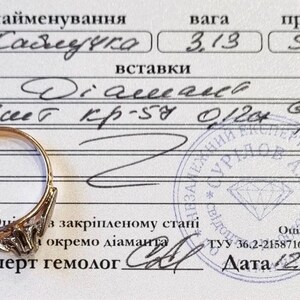 May include: A gold ring with a single diamond setting placed on a white certification document. The document features handwritten text including 3.13 weight, 583 purity, and diamond specifications, along with a circular blue ink stamp and a date.