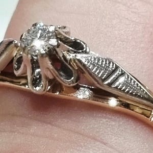 May include: A two-tone ring featuring a central round brilliant-cut diamond set in a white metal prong setting. The ring band is crafted from yellow gold, while the decorative shoulders are made of white metal with a textured, engraved leaf-like design.