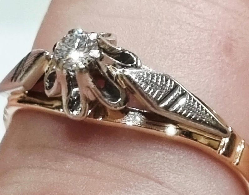 May include: A two-tone ring featuring a central round brilliant-cut diamond set in a white metal prong setting. The ring band is crafted from yellow gold, while the decorative shoulders are made of white metal with a textured, engraved leaf-like design.