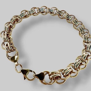Könnte beinhalten: Ein kreisförmiges Gliederarmband aus abwechselnd gold- und silberfarbenen Metallringen. Das Design zeigt ein sich wiederholendes Muster aus miteinander verbundenen, kreisförmigen Gliedern mit polierter Oberfläche und einem Karabinerverschluss.