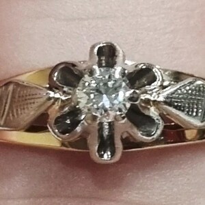 May include: A gold ring featuring a central round clear gemstone set within a six-petaled flower-shaped mount. The band includes two diamond-shaped sections on either side of the setting, each decorated with a fine cross-hatched engraved pattern.