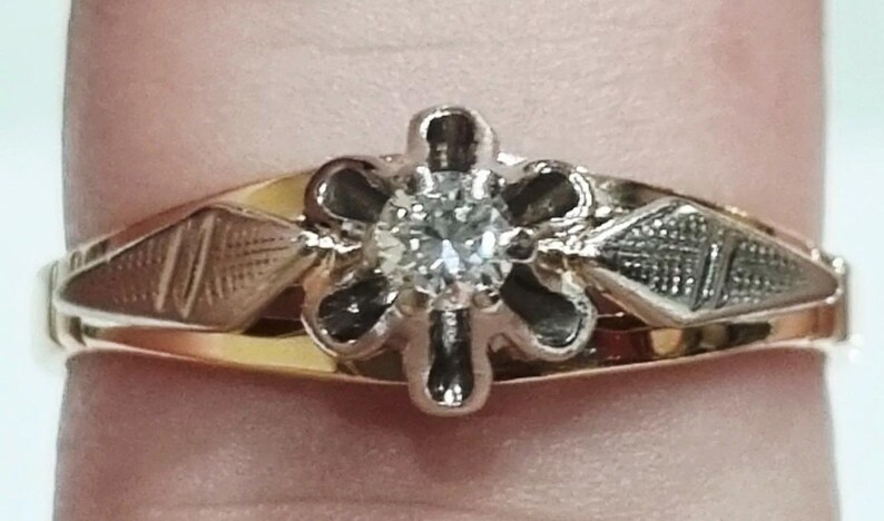 May include: A gold ring featuring a central round clear gemstone set within a six-petaled flower-shaped mount. The band includes two diamond-shaped sections on either side of the setting, each decorated with a fine cross-hatched engraved pattern.