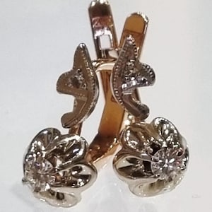 May include: A pair of rose gold earrings featuring a hinged lever back closure. Each earring has a dangling floral-shaped charm crafted from white gold, with a single clear round gemstone set in the center of each flower petal arrangement.