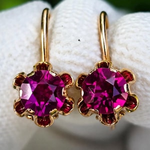 Peut inclure: Une paire de boucles d'oreilles en or avec des pierres rondes à facettes rose foncé, serties dans une monture en or en forme de fleur à six pétales. Chaque boucle d'oreille possède un crochet en or incurvé, avec une finition métallique polie.