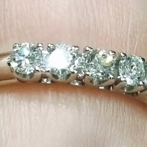 May include: A silver-toned metal ring featuring five round, clear, faceted stones set in a row along the top band. The stones are held in place by small metal prongs, creating a continuous, sparkling line across the front of the ring.