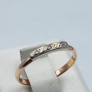 May include: A rose gold ring featuring a slender band and a rectangular top section in white metal. The top section is decorated with a textured, diagonal striped pattern and is set with three small, clear, round-cut stones arranged in a row.