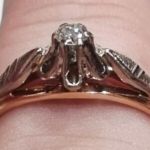 May include: A two-tone ring featuring a central clear gemstone set in a silver-colored prong setting, mounted on a gold-colored band. The shoulders of the ring display a textured, leaf-like pattern in a silver-colored metal finish.