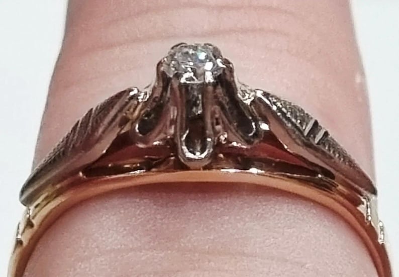 May include: A two-tone ring featuring a central clear gemstone set in a silver-colored prong setting, mounted on a gold-colored band. The shoulders of the ring display a textured, leaf-like pattern in a silver-colored metal finish.