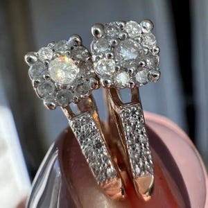 May include: A pair of rose gold earrings featuring square-shaped heads set with multiple round white stones in a cluster arrangement. The bands of the earrings are also encrusted with small white stones, creating a sparkling appearance.