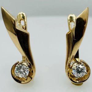 May include: A pair of gold earrings featuring a polished, curved, leaf-like metal design that tapers to a point at the top. Each earring is set with a single, round, brilliant-cut clear gemstone at the base, secured by a hinged lever-back closure.