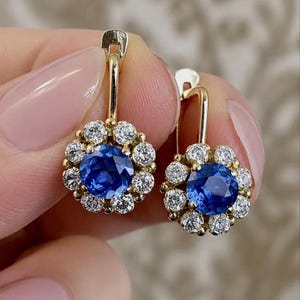 Pode incluir: Um par de brincos pendentes de ouro com uma pedra preciosa azul central de corte redondo rodeada por um halo de dez pequenas pedras brancas redondas e claras. Os brincos possuem um fecho de alavanca e um acabamento em ouro polido.
