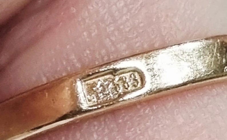 May include: A gold ring band featuring a rectangular hallmark stamp on the interior surface. The stamp displays a five-pointed star symbol positioned to the left of the number 585, indicating the gold purity of the metal band.