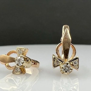 May include: A pair of rose gold earrings featuring a central round clear stone set within a decorative metal frame. Each earring has a pointed top element and two smaller clear stones on the sides, secured with a hinged lever back closure.