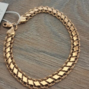 Könnte beinhalten: Ein roségoldfarbenes Armband mit einem komplexen, mehrschichtigen Kettenmuster aus sich wiederholenden rautenförmigen Gliedern, die durch kleinere kreisförmige Ringe verbunden sind. Das Armband verfügt über einen sicheren Karabinerverschluss.