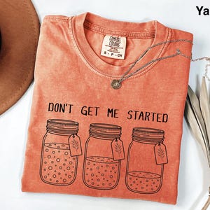 May include: A coral-colored t-shirt with the phrase "DON'T GET ME STARTED" printed in black. The design features three mason jars with tags. A silver necklace with a pendant is draped across the shirt.