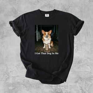 Op de afbeelding: Zwart t-shirt met een afbeelding van een corgi-hond die een bierblikje vasthoudt. De hond is bruin en wit, en het bierblikje heeft een etiket. De tekst "I Got That Dog In Me" staat eronder.