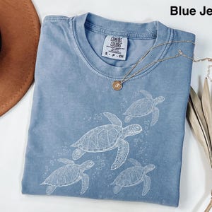 Könnte beinhalten: Ein blaues Jeans-T-Shirt mit einer weißen Grafik von vier schwimmenden Meeresschildkröten. Das Shirt hat einen Rundhalsausschnitt und kurze Ärmel. Eine goldene Halskette mit Anhänger liegt über dem Shirt.