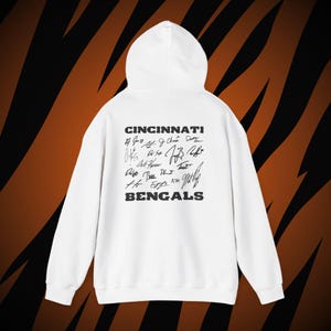 May include: A white hooded sweatshirt featuring the text CINCINNATI BENGALS in black block letters on the back. Between the words is a collection of black ink signatures arranged in a rectangular block. The hoodie has long sleeves and a hood.
