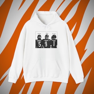 Op de afbeelding: Een wit sweatshirt met capuchon en lange mouwen met een zwarte grafische print op de borst. De afbeelding toont drie footballspelers van achteren met de namen Higgins, Burrow en Chase en de rugnummers 5, 9 en 1.