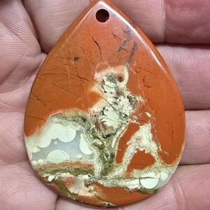 May include: Teardrop-shaped pendant with a hole at the top. The stone features a blend of orange, white, and green hues with natural patterns and cracks. The pendant is held in a hand.