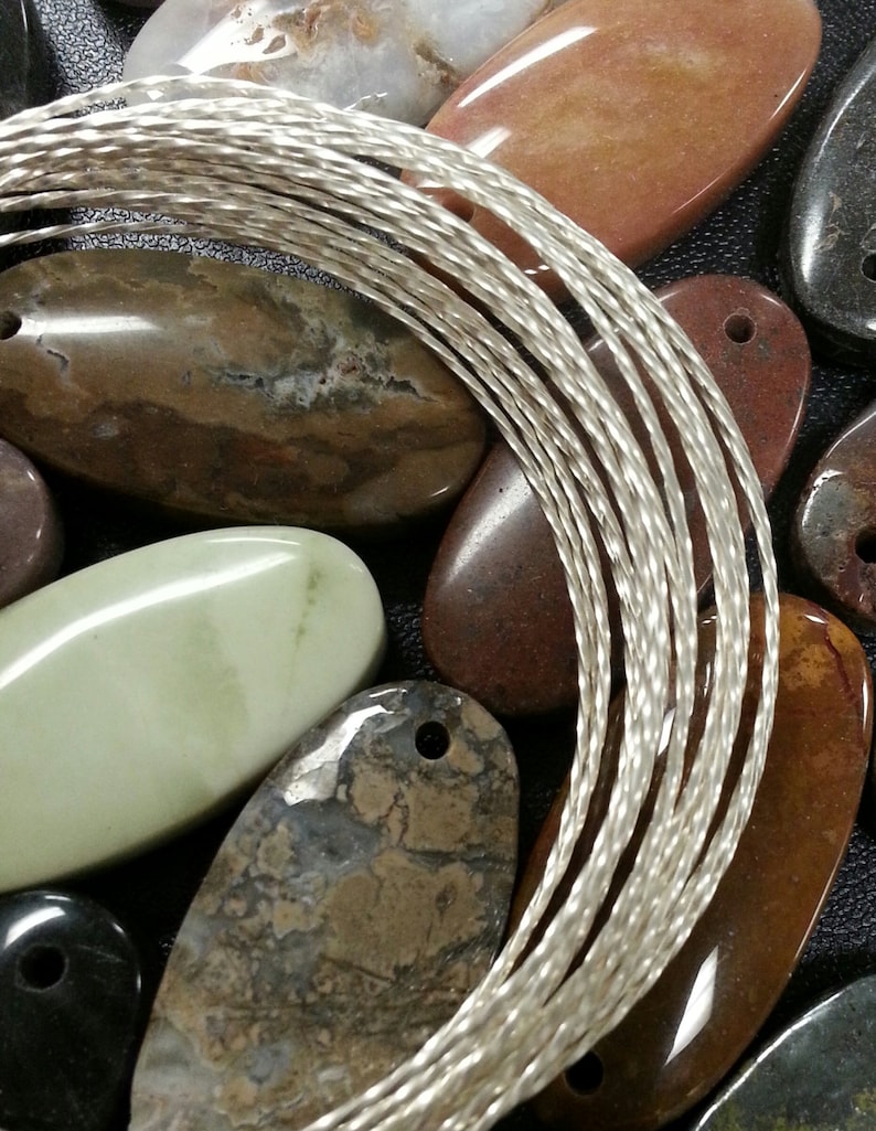 Puede incluir: Primer plano de un alambre trenzado plateado, con una variedad de piedras lisas de forma ovalada en el fondo. Las piedras son en tonos de marr&oacute;n, verde y negro.