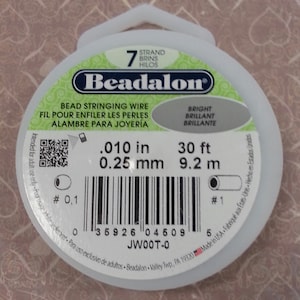 May include: A white spool of Beadalon bead stringing wire. The spool is labeled with the text "Beadalon", "Bead Stringing Wire", "Fil pour enfiler les perles", "Alambre para Joyeria", "Bright", "Brilliant", "Brillante", ".010 in", "30 ft", "0.25 mm", "9.2 m", "JWOOT-0", and "Made in USA".