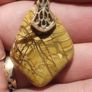 May include: A pendant with a geometric shape, featuring a patterned, tan-colored stone with brown veining. The pendant has a decorative, silver-colored bail. The jewelry is likely for necklaces or other accessories.