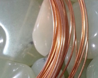May include: Close-up of coiled copper wire, showcasing its warm, reddish-brown color. The wire is in focus, with a blurred background of pale green, smooth stones. The image highlights the texture and sheen of the metal, suggesting craft supplies or jewelry making materials.