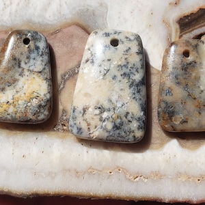 May include: Three natural stone pendants with a hole drilled through the top. The stones are a mix of white, gray, and brown with a mottled pattern.