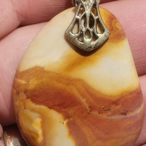 May include: A pendant featuring a polished, teardrop-shaped stone with swirling patterns of cream, orange, and brown. The stone is topped with a decorative silver-colored bail. The pendant is likely part of a jewelry collection.